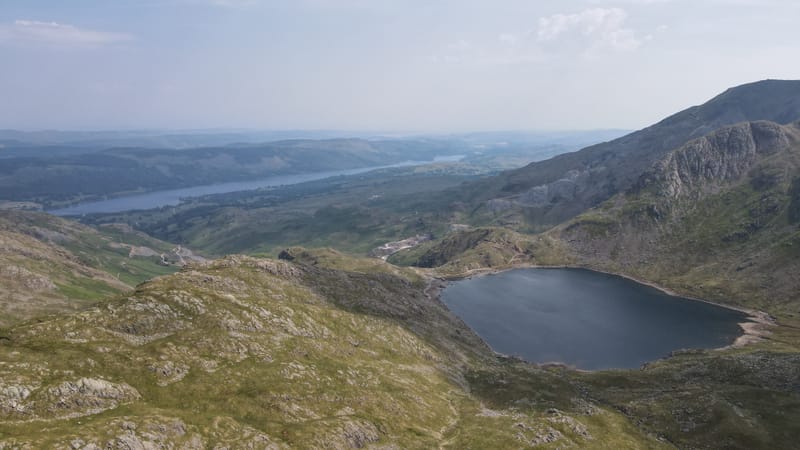 A dark mountain tarn below rocky crags, with a long lake in the valley beyond under a pale summer sky.