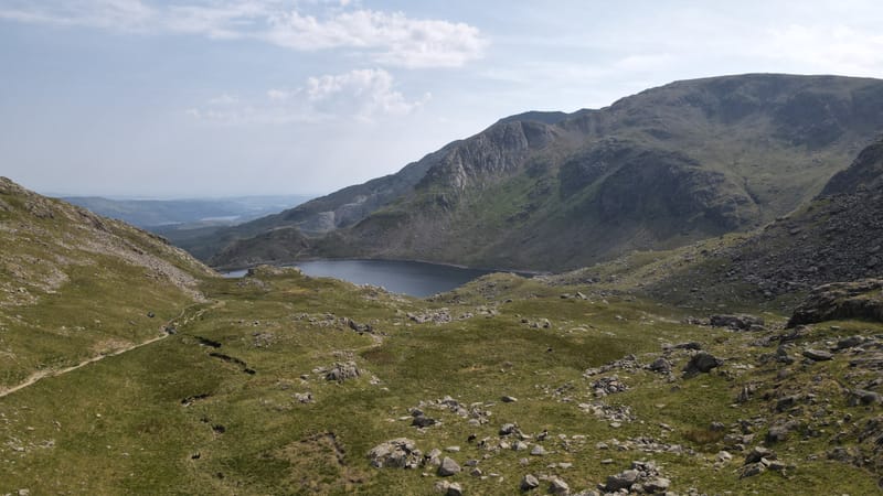 A mountain path through grass and boulders towards a tarn beneath a craggy ridge.