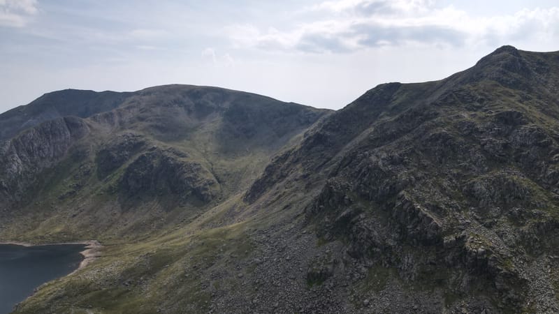 A rocky ridge rising above the edge of a tarn, under a hazy summer sky.
