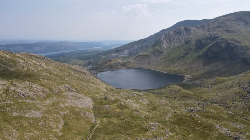 A mountain tarn in a grassy hollow under crags, with a lake and low hills in the distance.